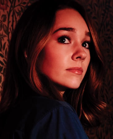 Holly Taylor headshot in dark blue top against black vine wallpaper looking over her shoulder under red lighting