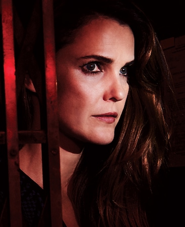 Keri Russell with long brown hair parted over left shoulder, staring up behind metal bars and illuminated with red light