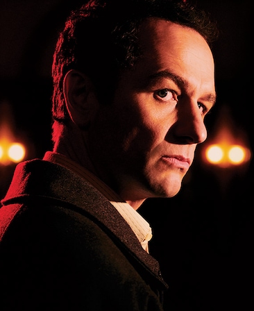 Matthew Rhys in black coat looking up over his right with half face illuminated at night with red lights in background