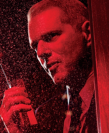 Noah Emmerich headshot holding walkie talkie in hand and looking up behind wet glass under red lighting