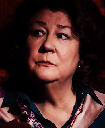 Margo Martindale headshot in pink silk coat with dark blue button down beneath, looking to her right with face illuminated