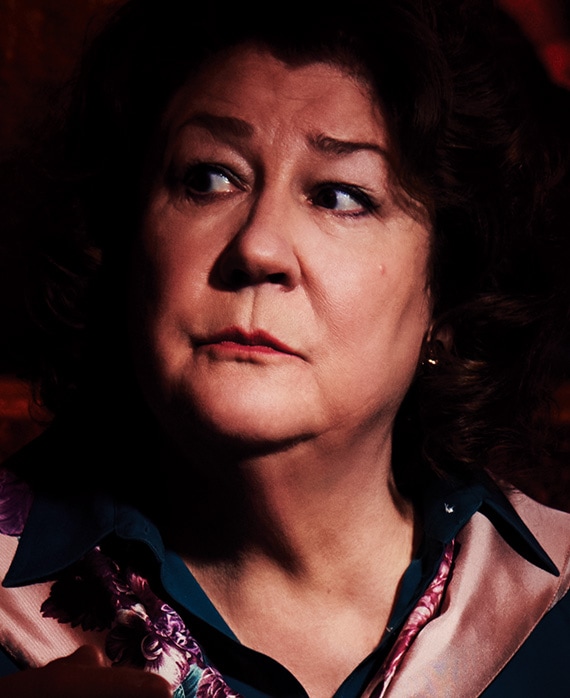 Margo Martindale headshot in pink silk coat with dark blue button down beneath, looking to her right with face illuminated