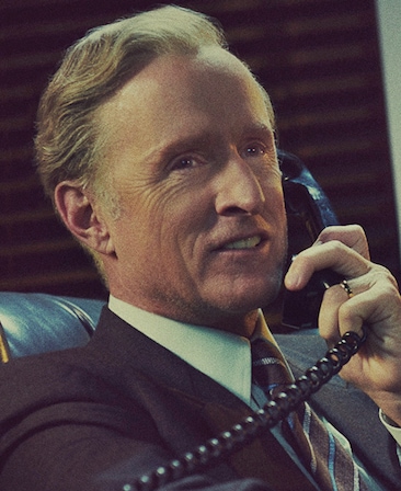 John Slattery headshot wearing a suit and holding a phone with a cord