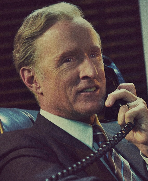 John Slattery headshot wearing a suit and holding a phone with a cord