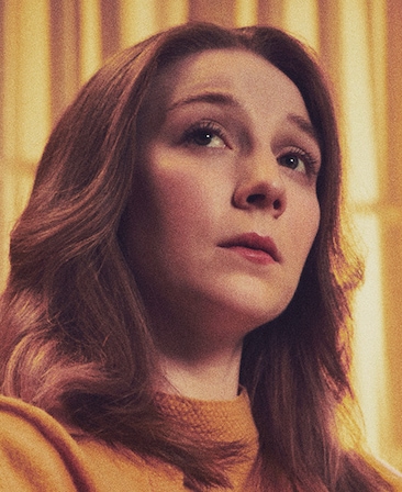 Kayli Carter headshot wearing an orange top in front of yellow curtains