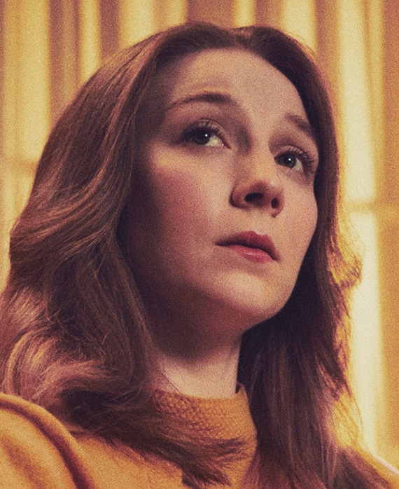 Kayli Carter headshot wearing an orange top in front of yellow curtains