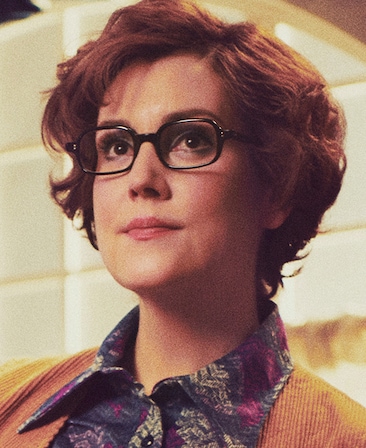 Melanie Lynskey headshot wearing a navy pattern blouse and orange jacket with black glasses