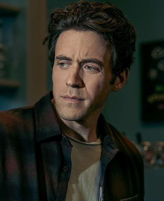 Ashley Zukerman Headshot wearing a white shirt and black jacket while looking down