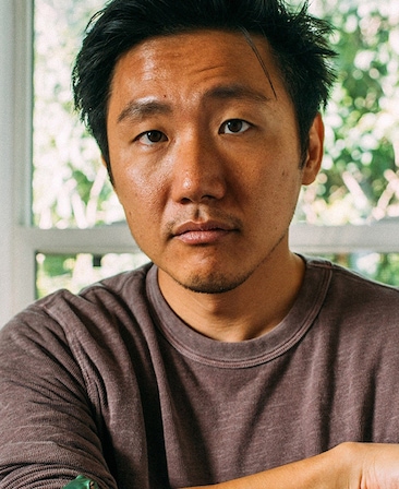 Hiro Murai headshot in dark maroon long sleeve shirt with light greenery in background