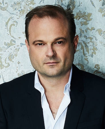 Brad Simpson headshot wearing a white shirt and black jacket with a patterned wall behind him