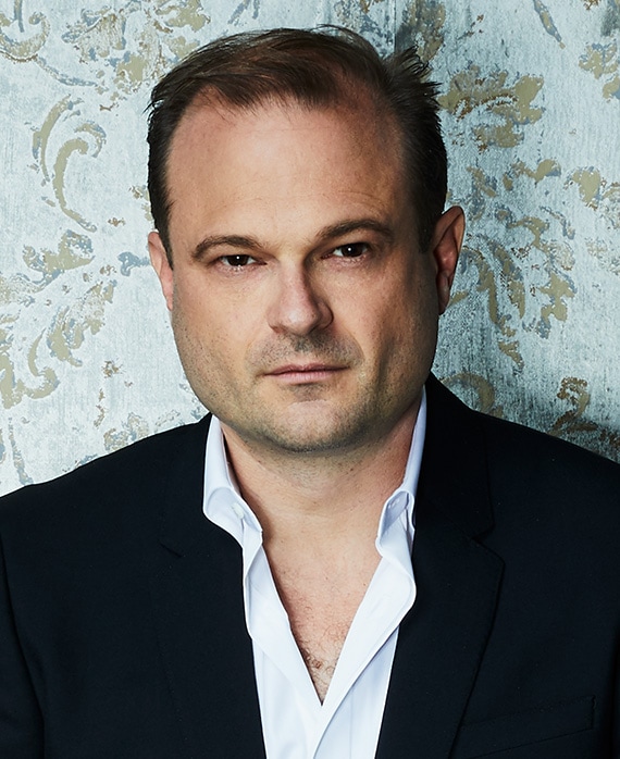 Brad Simpson headshot wearing a white shirt and black jacket with a patterned wall behind him
