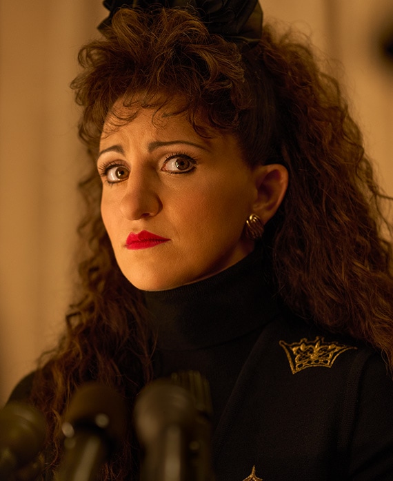 Headshot of Annaleigh Ashford as Paula Jones in black coat and bow in front of microphones from FX's American Crime Story