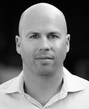 Black and white headshot of Justin Falvey in checkered button down against black and white background