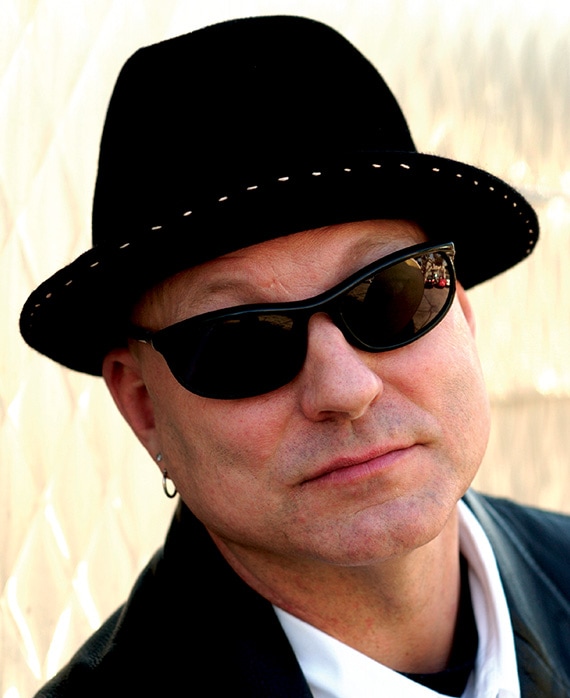 Charles H Eglee headshot wearing a black fedora with white stitching and black sunglasses, with an earring in his ear