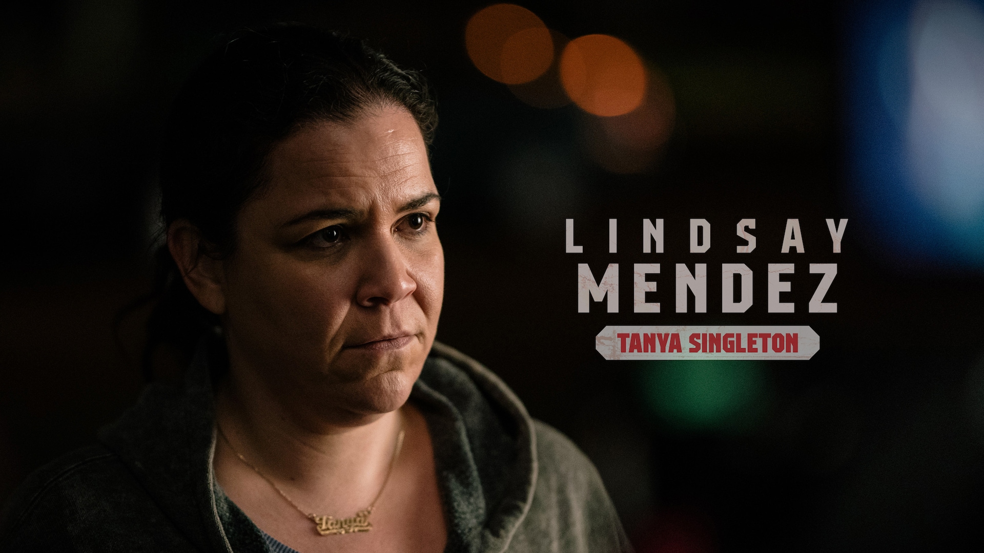 Lindsay Mendez as Tanya Singleton wearing a green hoodie and gold necklace outdoors with blurry lights behind her.