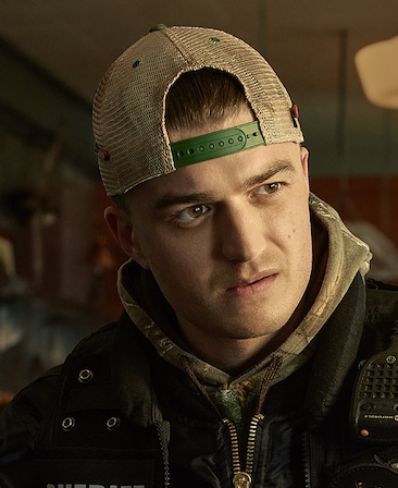 Joe Keery headshot wearing a backwards trucker hat with a hoodie tucked under his jacket