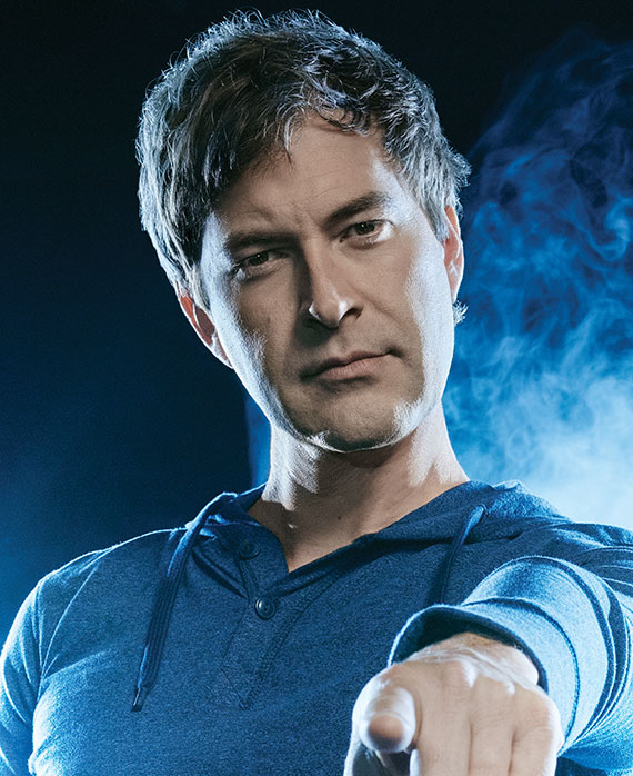 Mark Duplass headshot in blue henley sweatshirt, pointing at front with left hand and smoke in background with bright lights