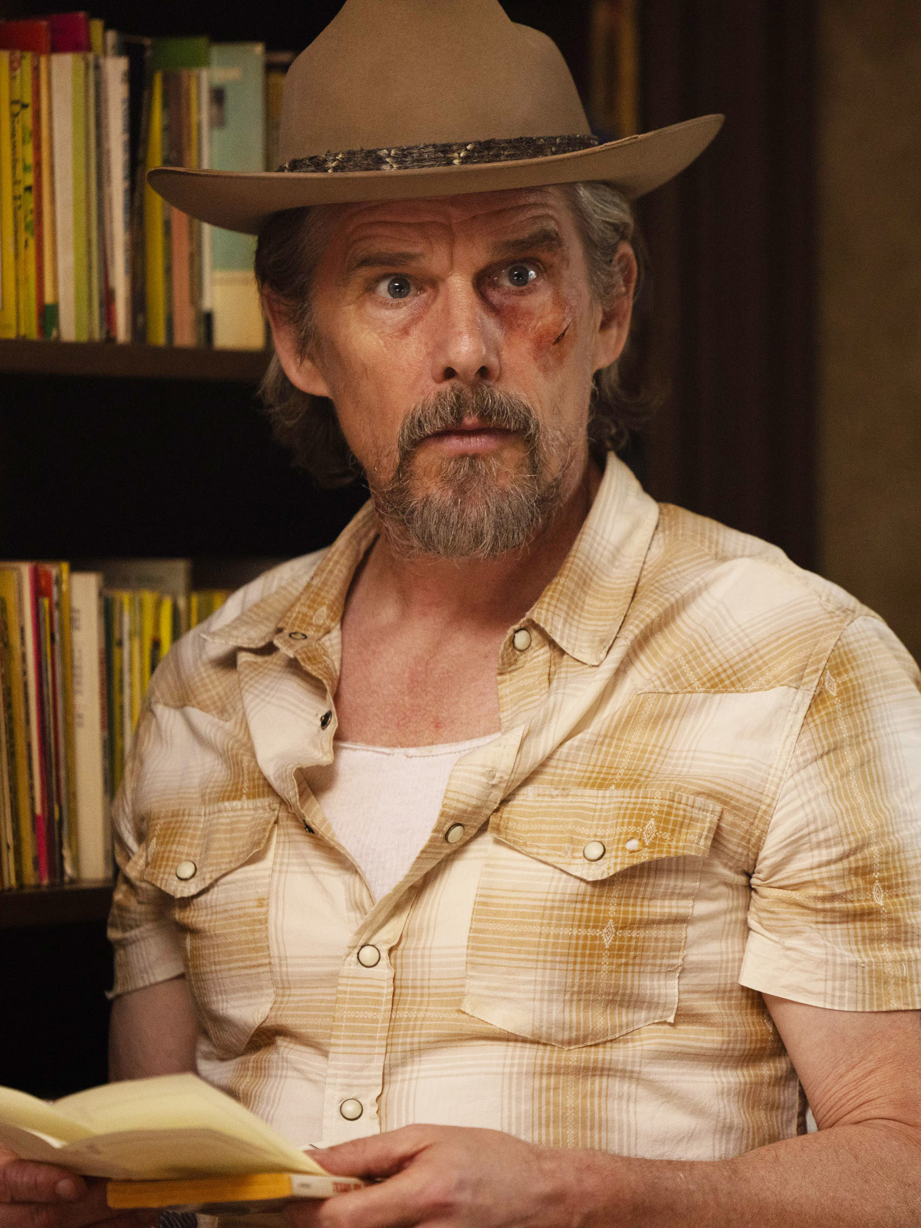 Ethan Hawke wearing a plaid shirt and a brown hat while holding a pamphlet