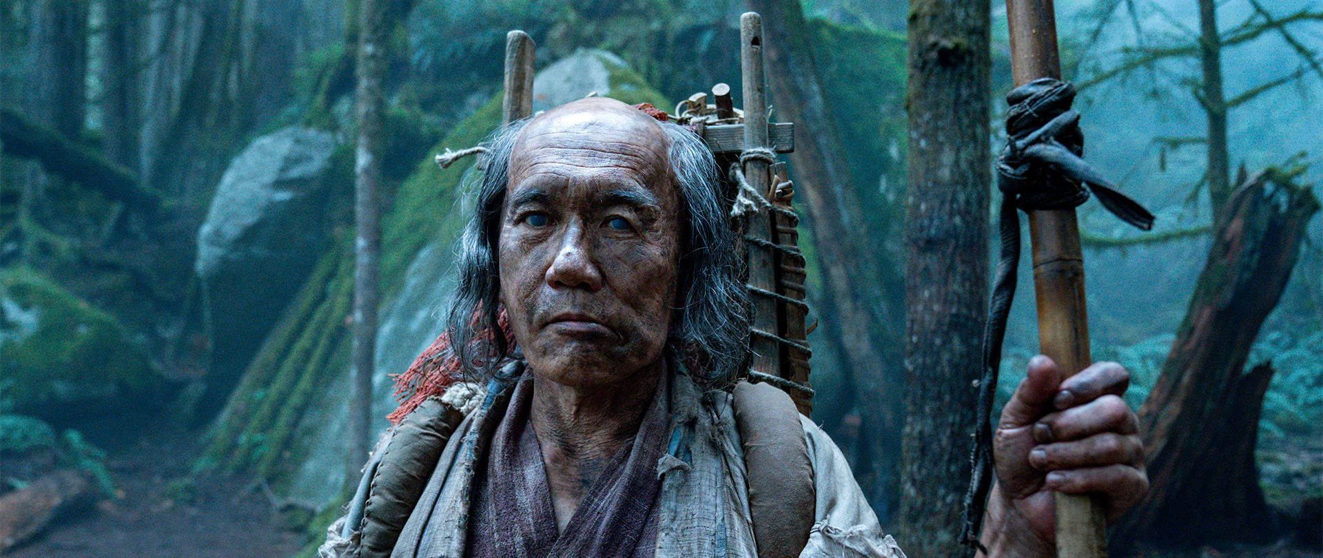 The blind beggar stands amid the forested set of FX's Shogun