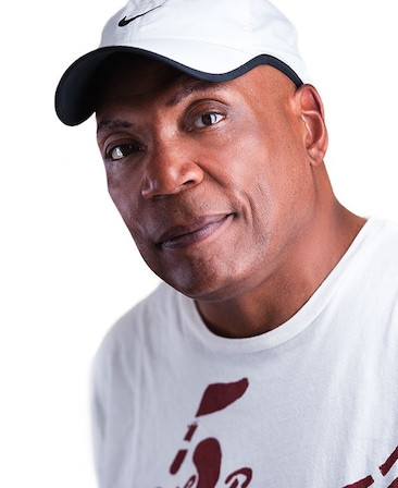 Paris Barclay headshot in white graphic t shirt with dark red logo and white and black Nike cap