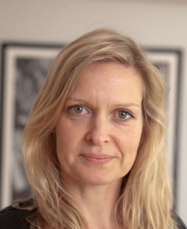 Kate Crowe headshot in natural lighting with framed black and white picture in background hanging on white wall