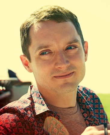 Elijah Wood headshot wearing a red shirt and patterned button up