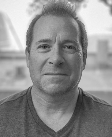 Black and white headshot of executive producer Jeff Luini, wearing a v-neck shirt, outdoors.