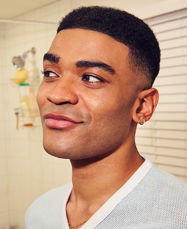 Headshot of Owen Thiele as Anton in FX's Adults wearing a mesh white v-neck shirt, wearing dainty gold jewelry.
