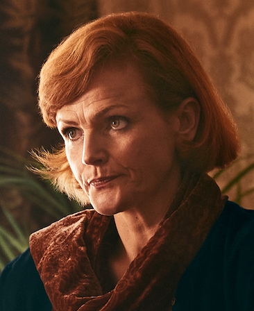 Maxine Peake as Older Delours Price, wearing a black coat and brown velvet scarf.