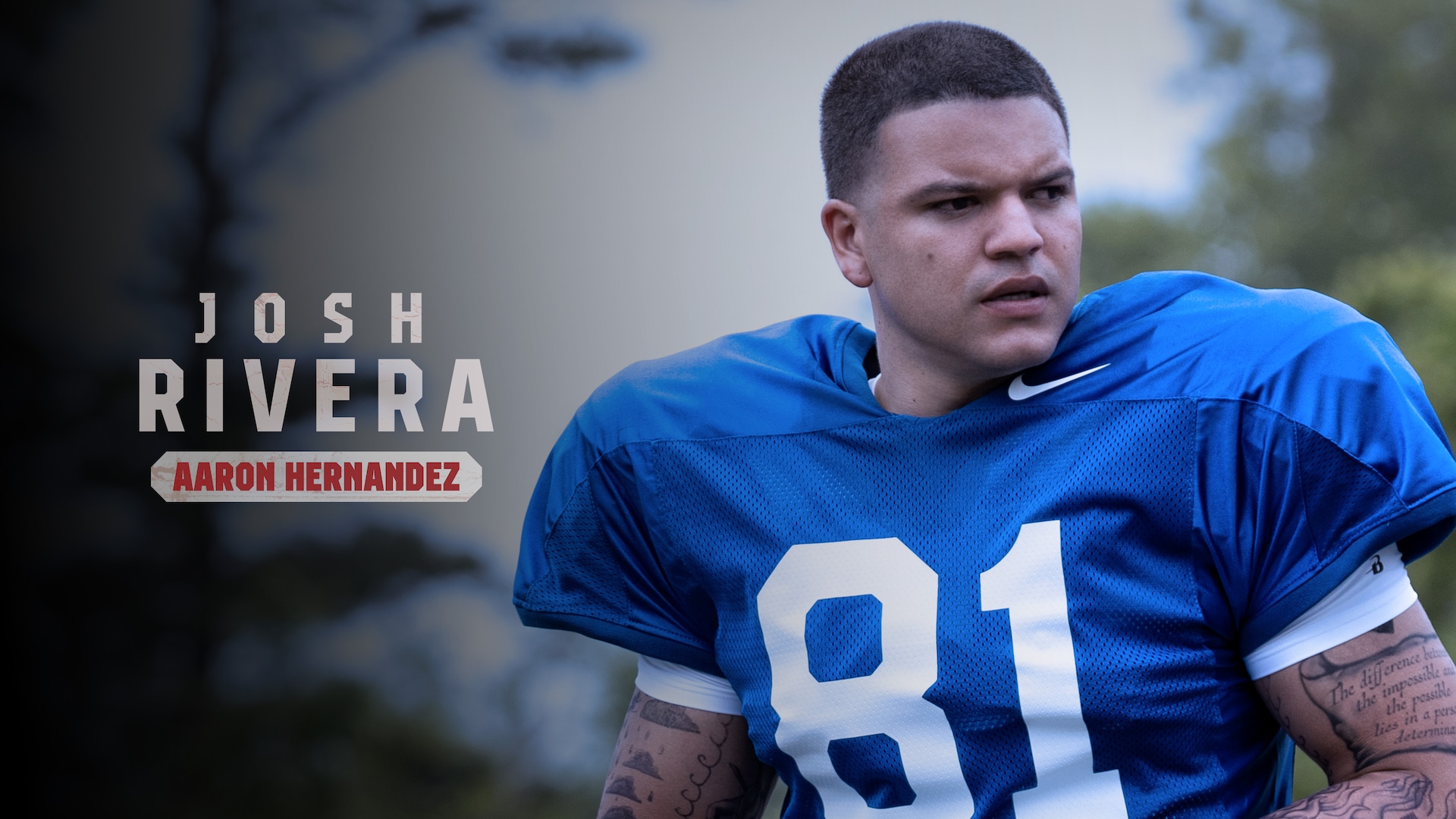 Josh Rivera as Aaron Hernandez wearing a blue football jersey, outdoors.