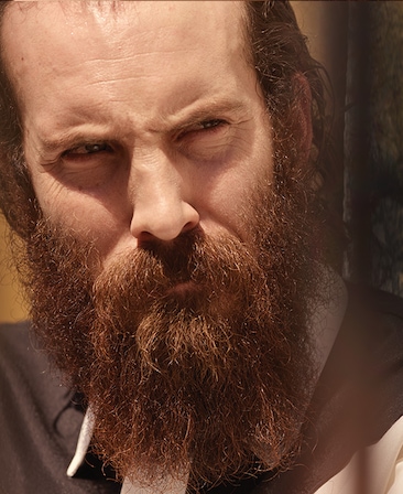 Thomas Wright headshot in dark brown and white top, looking to the side with bright sun shining down on his face